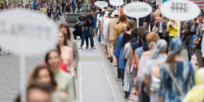 A camera team standing between two rows of models walking across a public space in front of an audience.