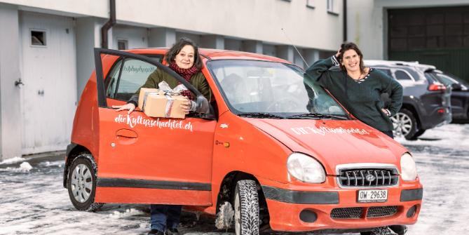 Die zwei Gründerinnen von DIE Kulturschachtel stehen neben ihrem beschrifteten Auto, eine hält ein Päckli in der Hand